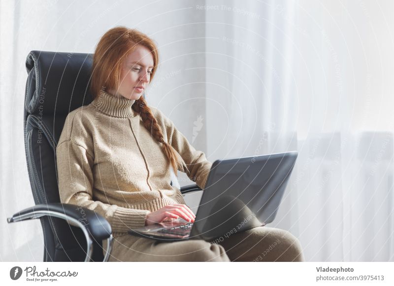 Junge kaukasische Frau, die in einem Ledersessel sitzt und einen Laptop benutzt, um von zu Hause aus zu arbeiten, Freiberufler, soziale Distanzierung und Fernarbeit.