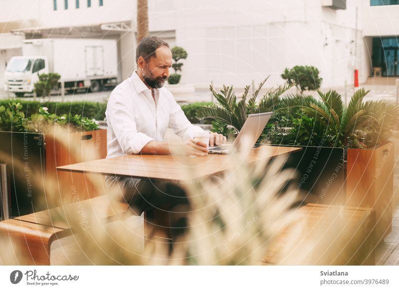 Erwachsener männlicher Geschäftsmann sitzt auf der Straße in einem Café, trinkt Kaffee und arbeitet an einem Laptop tausendjährig Mann Freiberufler Smartphone