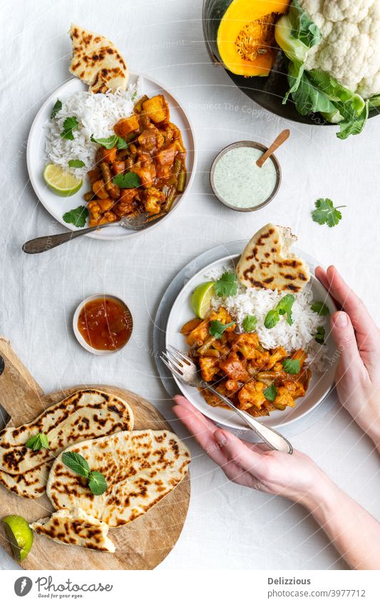 Draufsicht auf ein vegetarisches, veganes indisches Balti-Curry mit Blumenkohl und Kürbis, serviert mit Raita, Naan-Brot und Mango-Chutney und Reis, weibliche Hände halten einen Teller, weißer Hintergrund