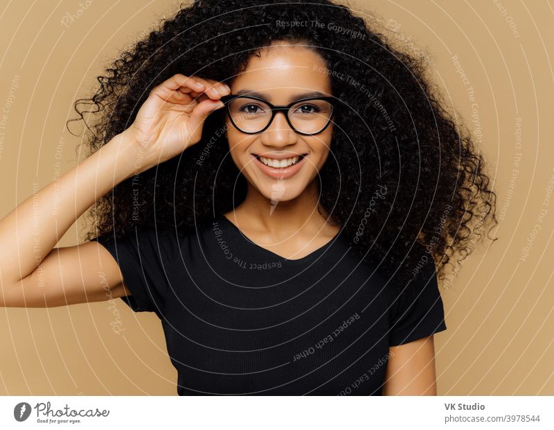 Lovely junge afroamerikanische Frau grinst in die Kamera, hält Hand auf Rand der Brille, froh, gute Nachrichten zu hören, in lässigen schwarzen T-Shirt gekleidet, isoliert über braunen Hintergrund. Glück Konzept