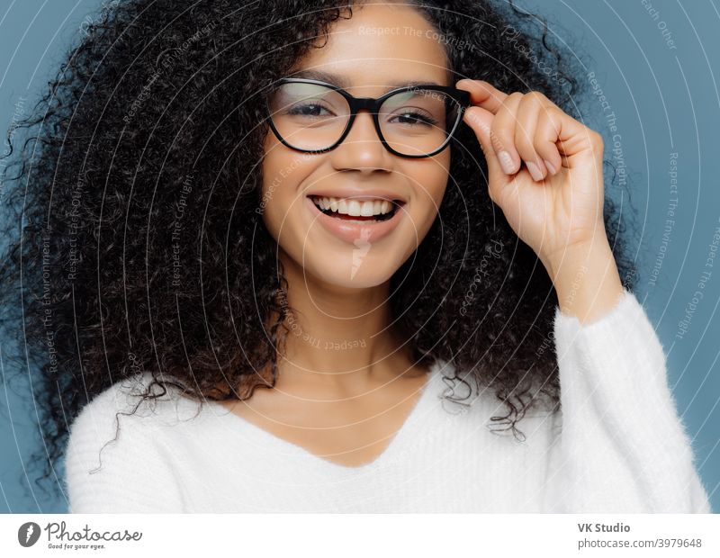 Cropped Schuss von gut aussehende Frau trägt optische Brille, lächelt breit, zeigt weiße perfekte Zähne, gesunde dunkle Haut, in lässigen Pullover gekleidet, isoliert über blauen Hintergrund. Feminität Konzept