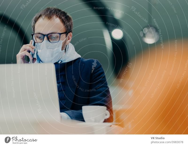 Geschäftsmann arbeitet von der Cafeteria aus, konzentriert auf das Display des Laptops, führt ein Telefongespräch, trägt eine medizinische Maske im Gesicht als Schutz vor Grippe oder Coronavirus-Epidemie. Intelligentes Arbeiten