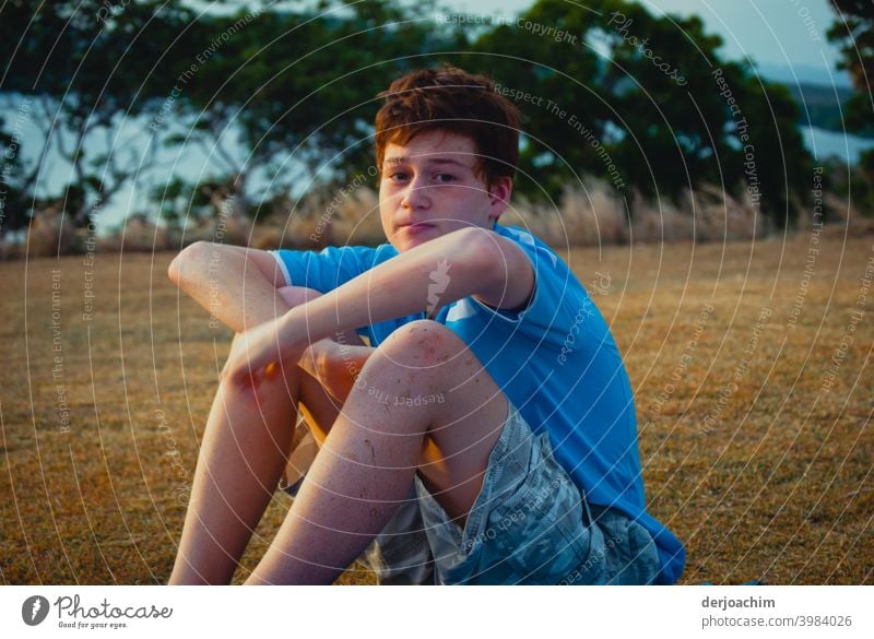 Ein Junge sitzt mit verschränkten Armen auf dem Boden. Im Hintergrund sind Büsche und blau schimmertes Wasser zu sehen. Er blickt in die Kamera. Junger Mann