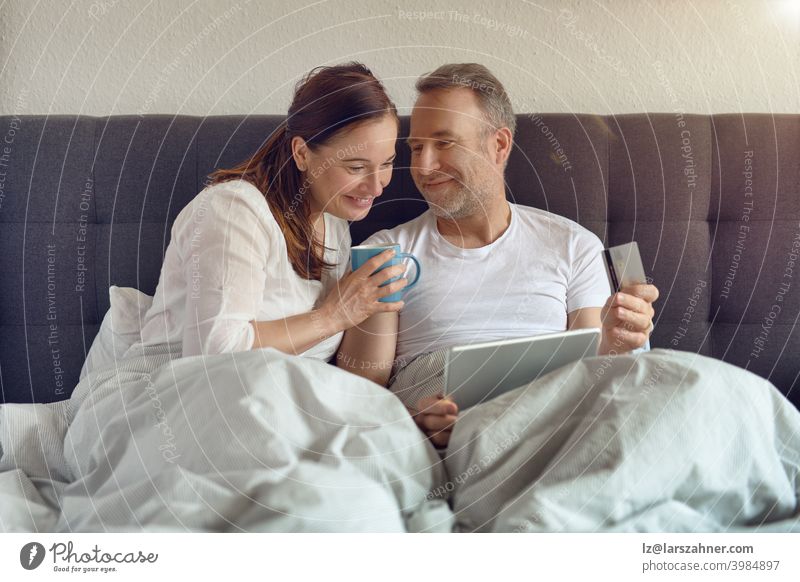 Glückliches Paar mittleren Alters, das morgens im Bett sitzt, Kaffee trinkt und mit einem digitalen Tablet Online-Einkäufe tätigt Sofa Möbel Familie Raum