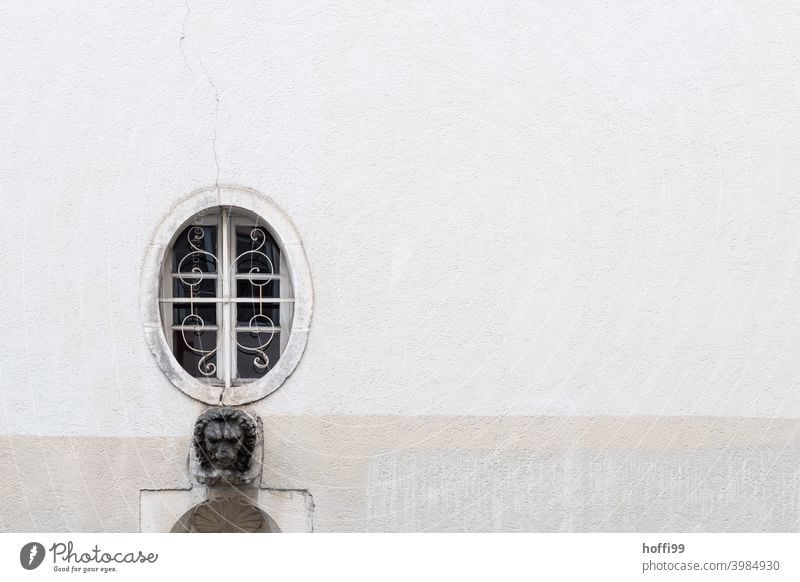 ein vergittertes ovales Fenster mit Löwenkopf auf grauer Wand Oval rund vergitterte Fenster Altstadthaus rundes fenster außergewöhnlich minimalistisch