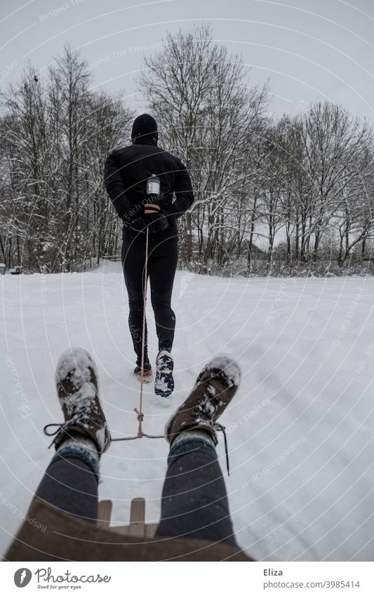 Ein Mann joggt im Winter durch den Schnee und zieht dabei eine Person auf einem Schlitten hinter sich her joggen ziehen Sport sportlich Menschen Paar draußen