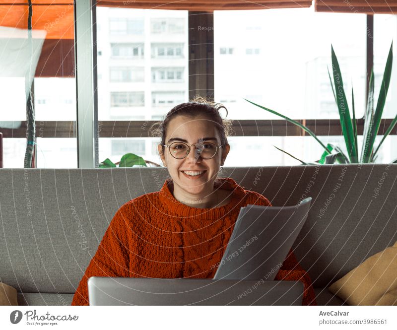 Eine junge Frau mit Brille arbeitet an seinem Laptop auf der Couch zu Hause, während sie in die Kamera lächelt Dokumente horizontal Job lebend entspannt