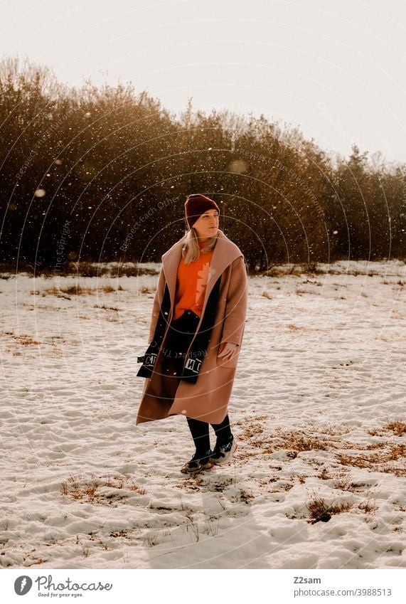 Modisch gekleidete Junge Frau geht bei leichtem Schneefall spazieren schneien winterwonderland frau junge frau mantel spaziergang schnee sonne licht natur