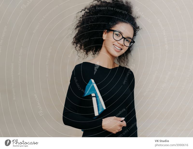 Positive lockige afroamerikanische Studentin trägt Notizblock und Tagebuch, trägt schwarze Kleidung, transparente Brille, genießt das Studium in der Universität, isoliert auf grauem Hintergrund. Nette Frau mit Notizbuch
