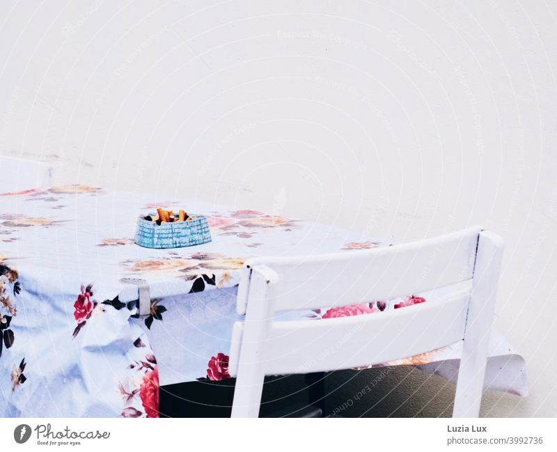 Raucherecke oder ein übervoller, blauer Aschenbecher auf einem Tisch mit altmodischer Wachsdecke, Blumenmuster und sommerliches Weiß Raucherplatz
