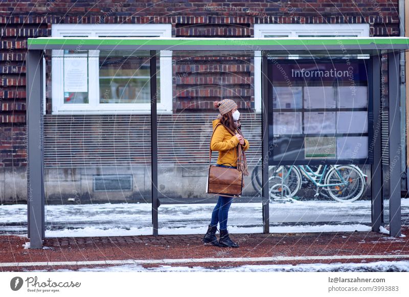 Brünette Frau mittleren Alters, die an einer Bushaltestelle steht, eine schützende Gesichtsmaske aufgrund des Coronavirus trägt und auf ihren Bus wartet, der sie zur Arbeit bringt