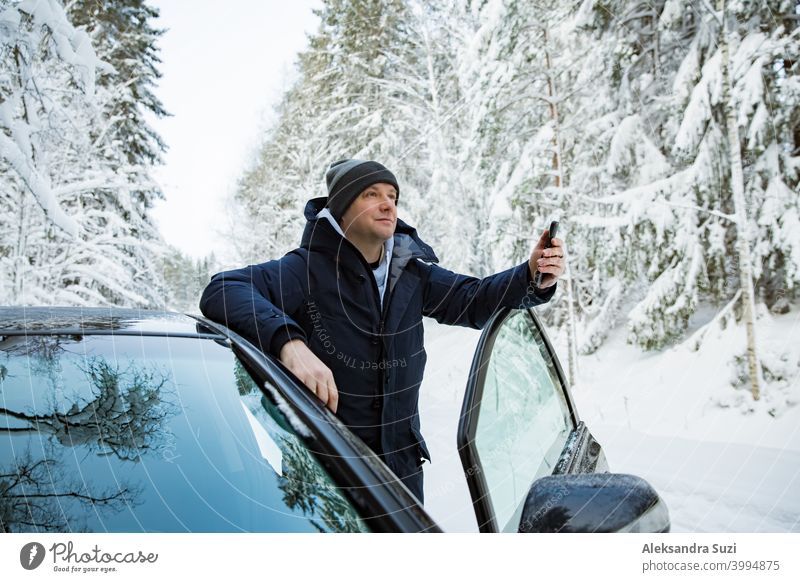 Mann in warmer Winterkleidung steht am Auto, mit Telefon. Snowy Winter Landstraße, Auto mit Eis bedeckt, Schöne Wald unter dem Schnee. Hilfsbereitschaft Anruf