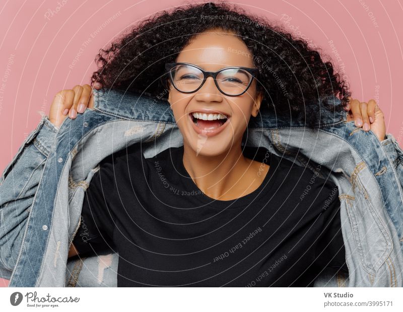 Isolierte Aufnahme von lockigen fröhliche Frau versucht auf neue Jeansjacke, trägt optische Brille, lächelt breit, hat perfekte Stimmung, posiert gegen rosa Wand. Positive Emotionen. Afro-Frau in stilvoller Kleidung