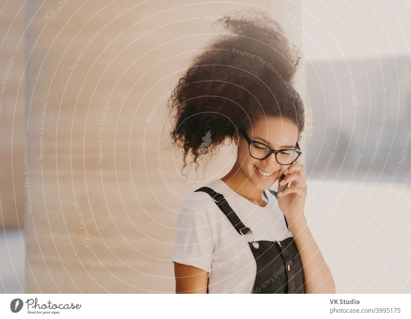 Millennial-Afro-Frau spricht über Handy, nutzt Roaming-Verbindung, schaut nach unten, genießt angenehme Unterhaltung, trägt weißes T-Shirt und Latzhose, ruht sich im Freien aus, telefoniert über App, hat gute Laune
