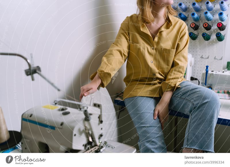 Gesichtslose Aufnahme einer jungen Frau in gelbem Hemd, die in ihrer Nähwerkstatt vor den bunten Fäden sitzt Näherin Maschine Textil Schneider Overlock Gewinde