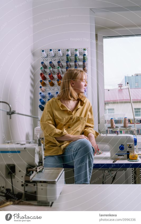 Junge Frau in gelbem Hemd sitzt in ihrer Nähwerkstatt vor den bunten Fäden und Nähmaschinen Näherin Maschine Textil Schneider Overlock Gewinde Werkstatt Beruf