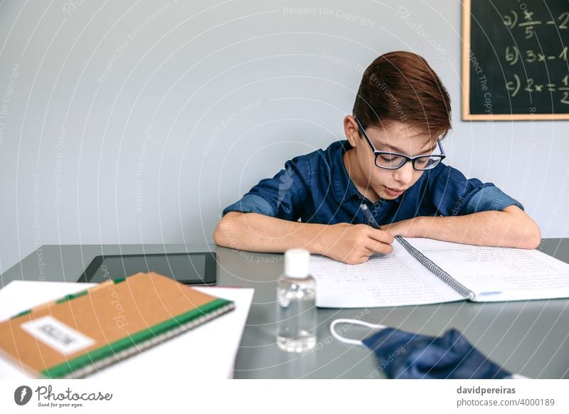 Junge schreibt in der Schule in sein Notizbuch Klassenraum Maske auf dem Schreibtisch Handdesinfektionsmittel Coronavirus Sicherheit schreibend Virus Menschen
