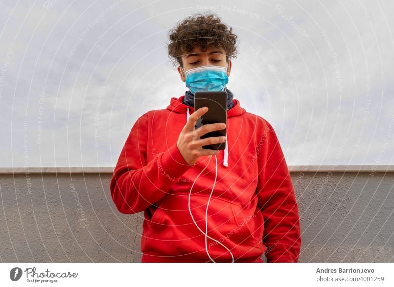 Junger attraktiver Mann mit lockigem Haar und rotem Sweatshirt, der auf der Terrasse des Hauses das Handy benutzt und eine Maske trägt, um sich vor dem Coronavirus zu schützen