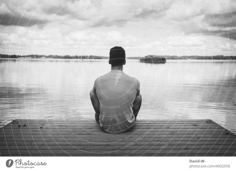 Mann sitzt einsam und alleine auf einem Steg am See und genießt die Ruhe Erholung Stille Abstand nachdenklich sitzen genießen Einsamkeit Rückansicht Seeufer