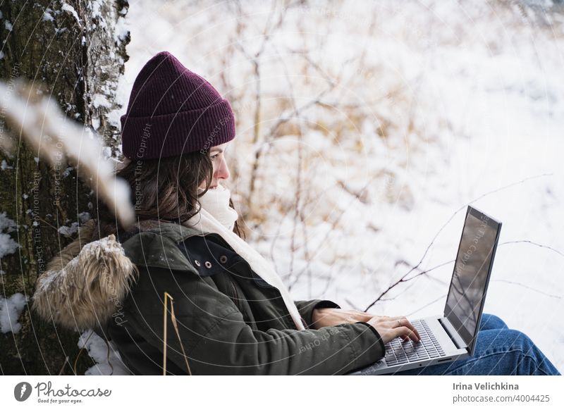 Junges Mädchen, Frau arbeitet in Laptop. Walking in schönen Winterwald unter Bäumen, Tannen, mit Schnee bedeckt. Magnificent Natur und Ansichten. Modisches Bild, Kleidung, Parka, Hut, Handschuhe, Blue Jeans