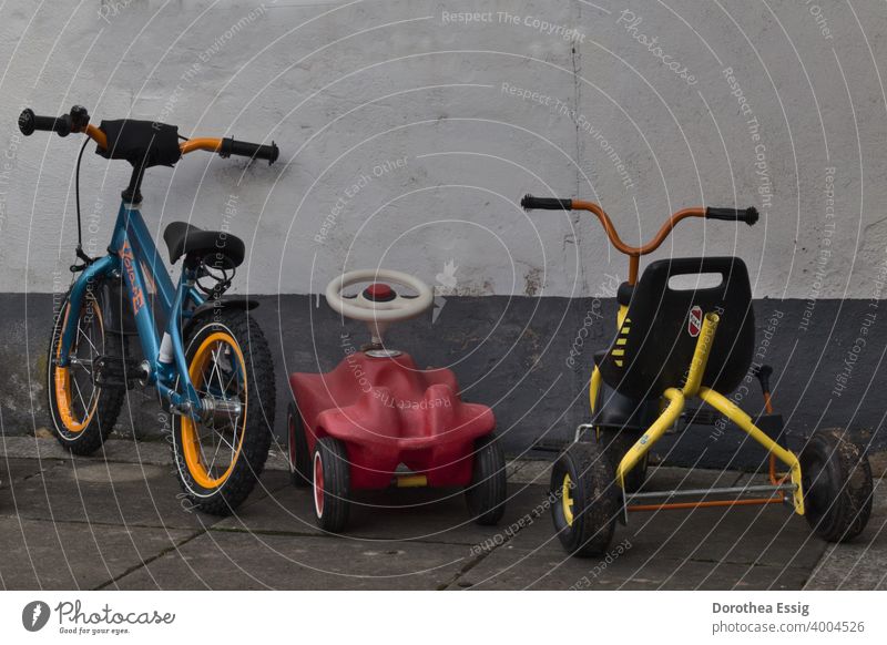 Ein Kinderfahrrad, ein Rutscheauto, ein Dreirad an einer Mauer abgestellt Kita Kinderfahrzeuge Fahrrad Bobbycar geparkt Spielzeug draußen Außenaufnahme Farbfoto