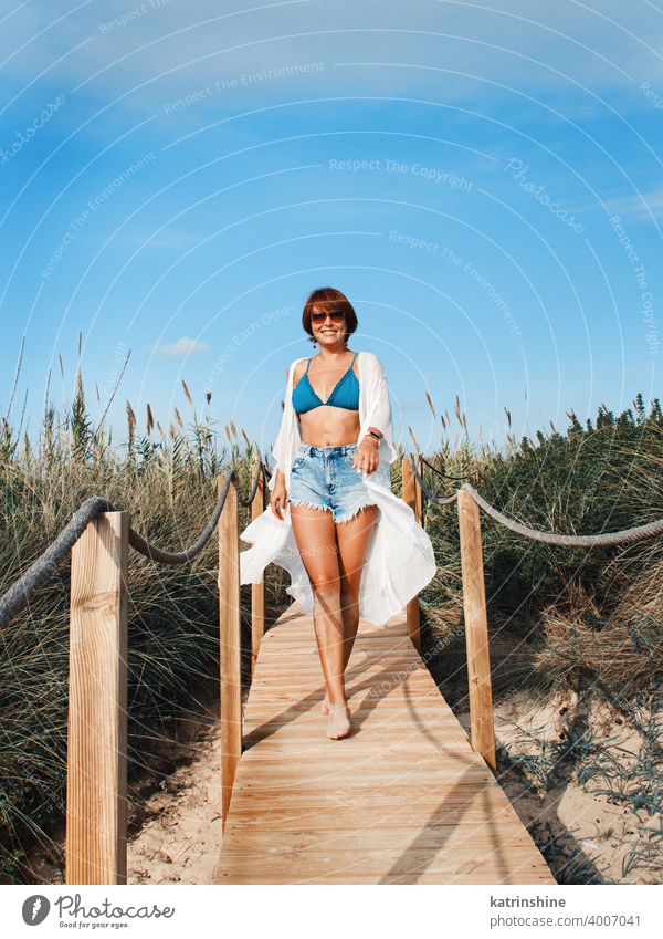 Junge Frau in Bedeckung und Jeans-Shorts, die zwischen den Dünen spazieren geht. jung zudecken Kimono weiß Lächeln allein blau türkis Jeanshose laufen Strand