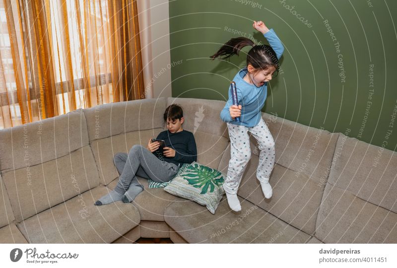 Mädchen springt und spielt auf dem Sofa, während sein Bruder auf dem Tablet sitzend schaut springend Liege Spielen Homewear Teenager Tablette Spaß Schrei