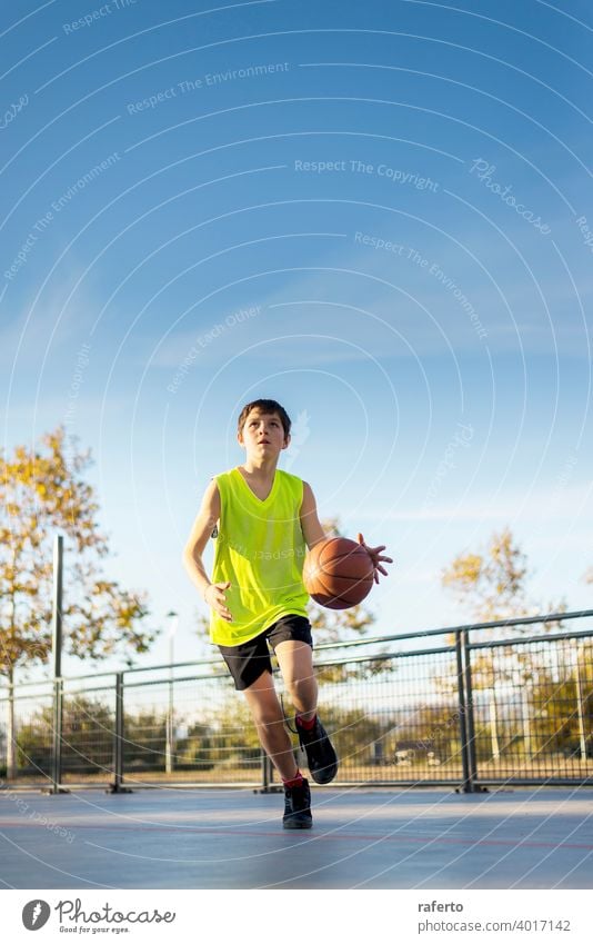 Süßer Junge in gelbem Hemd spielt Basketball auf dem Spielplatz der Stadt. Spieler Teenager männlich Ball Training Korb außerhalb springend Sport im Freien