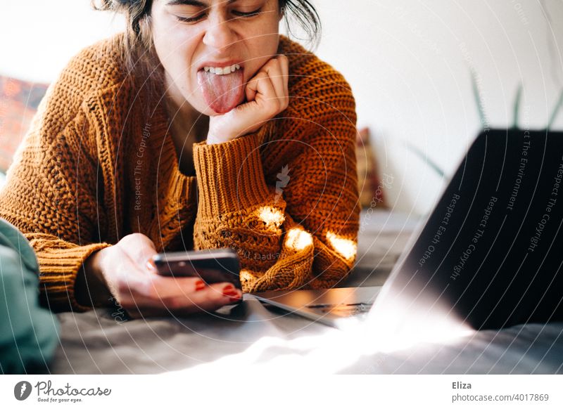 Eine Frau liegt zuhause neben ihrem Notebook im Bett und streckt ihrem Smartphone die Zunge raus. Genervt vom Home Office. Homeoffice Laptop arbeiten surfen