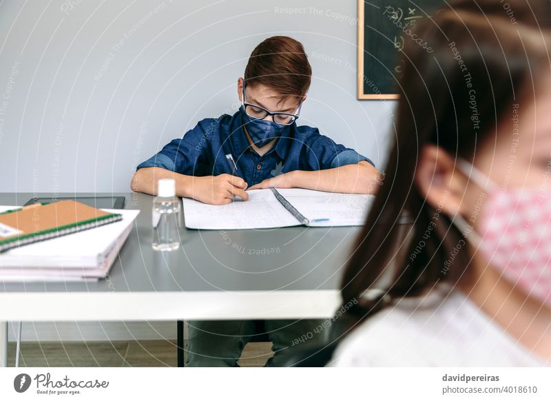 Junge mit Gesichtsmaske beim Schreiben in der Schule Schüler Klassenraum neue Normale Coronavirus Sicherheit schreibend Virus Menschen Seuche Mundschutz Bildung