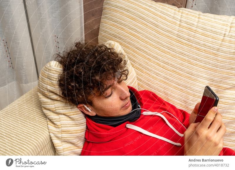 Junger attraktiver Mann mit lockigem Haar und rotem Sweatshirt, der im heimischen Wohnzimmer ein Handy benutzt und eine Maske trägt, um sich vor dem Coronavirus zu schützen
