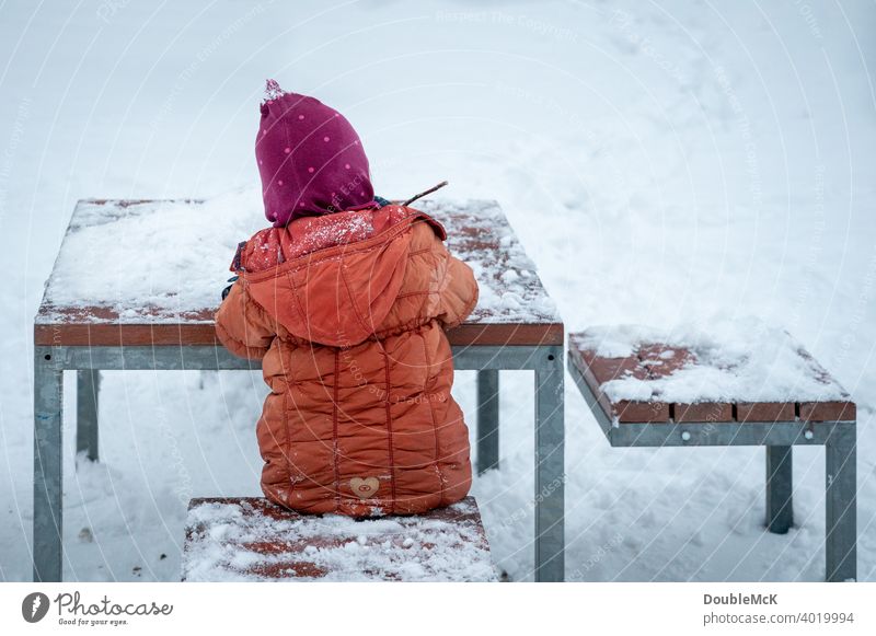 Ein Kind sitzt draußen am verschneiten Tisch im Schnee und spielt Tag Außenaufnahme natürlich Leben Freude kalt kalte jahreszeit weggucken Winter Kälte 1