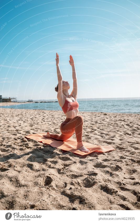 Junge Frau in orangefarbenem Oberteil und Leggins beim Üben der Sichel-Lunge-Pose (Anjaneyasana) am Strand am Morgen Übung Yoga jung praktizieren im Freien
