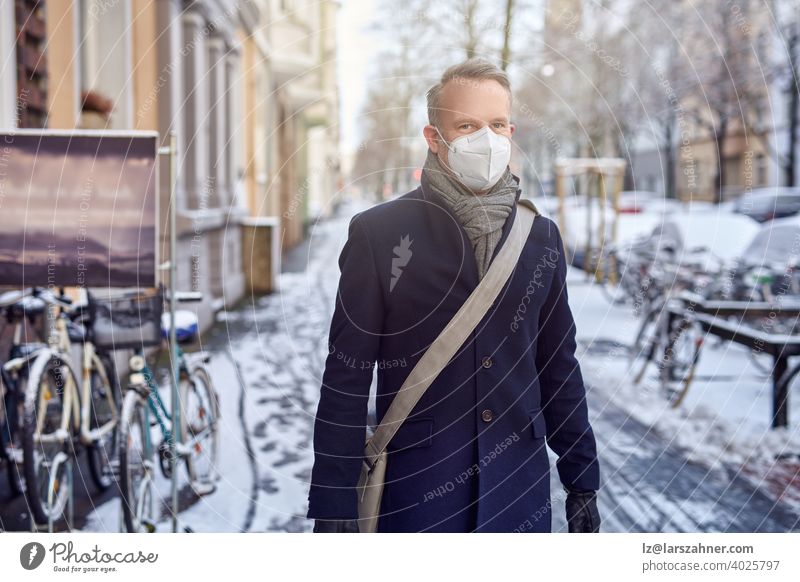 Mann mit einer schützenden chirurgischen Gesichtsmaske während der Covid-19- oder Coronavirus-Pandemie und Wintermantel mit Ledertasche über der Schulter, der eine verschneite städtische Straße mit geparkten Fahrrädern in Nahaufnahme entlanggeht und zur Seite schaut