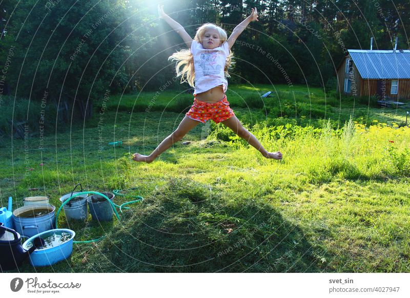 Barfuß Kindheit auf dem Dorf. Porträt eines kleinen blonden Mädchens, das in der Luft über einen kleinen Stapel frisch geschnittenen Grases auf einem grünen, sonnigen Rasen vor dem Hintergrund eines Waldes und eines hölzernen Badehauses springt.