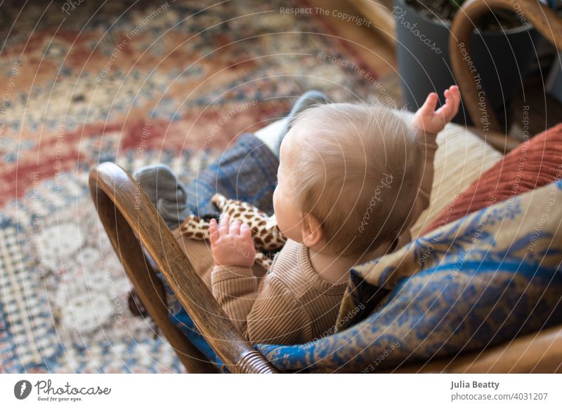 Junges Baby sitzt in Vintage Bambus Rattan Stuhl hält Spielzeug; bunten böhmischen Teppich und Dekor Kind Weide retro altehrwürdig Boho Bohemien gekrümmt
