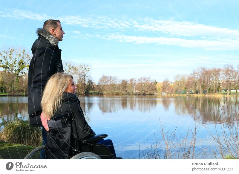 Konzept einer behinderten Person. Eine Frau im Rollstuhl mit einem Mann, der neben ihr steht, draußen in der Natur vor einem See Erwachsener Kontemplation Paar