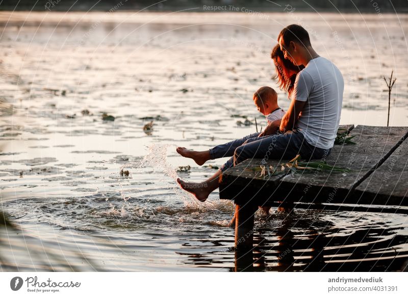 Junge Eltern und ihr kleiner Sohn sitzen auf dem Holzsteg in der Nähe des Sees, bei Sonnenuntergang am Sommertag. Familie Mutter jung Herbst Vater fallen