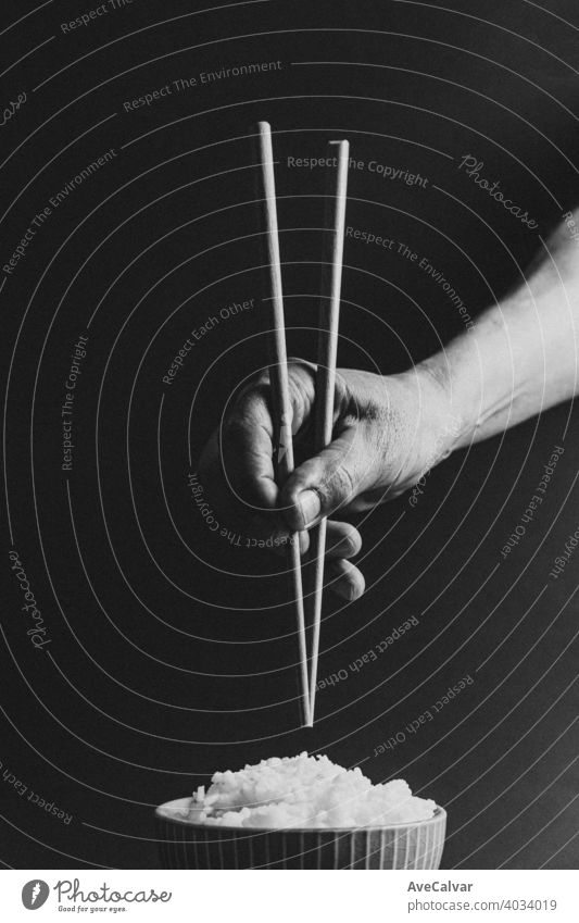 Minimalistische alte Hände greifen japanische Essstäbchen über eine Schüssel Reis Konzept Schuss auf filmische Töne über einen schwarzen Hintergrund in Schwarz und Weiß