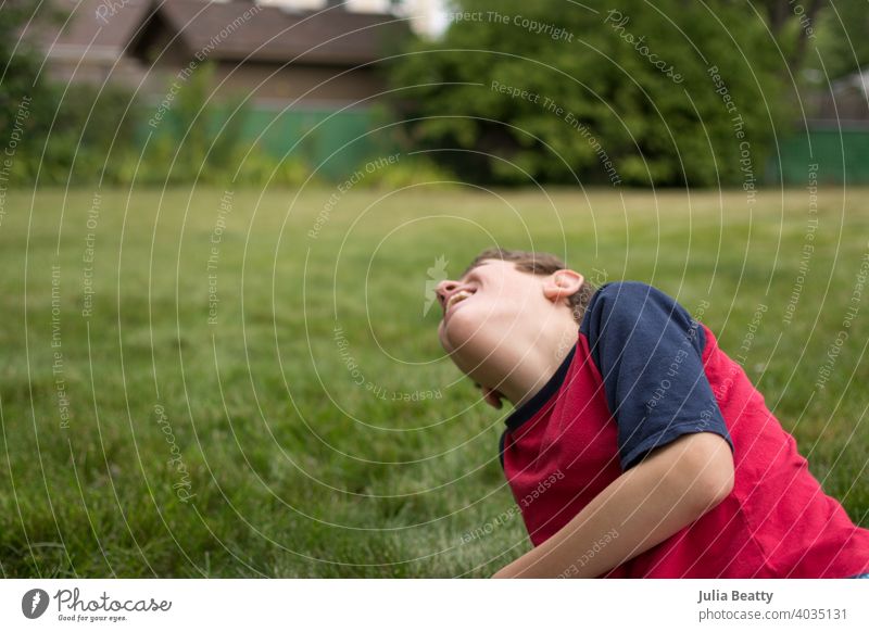 Junge mit Autismus lachend beim Sitzen im Gras in seinem Hinterhof; Sommerzeit Spaß besondere Bedürfnisse Kommunizieren Mitteilung Autismus-Bewusstsein