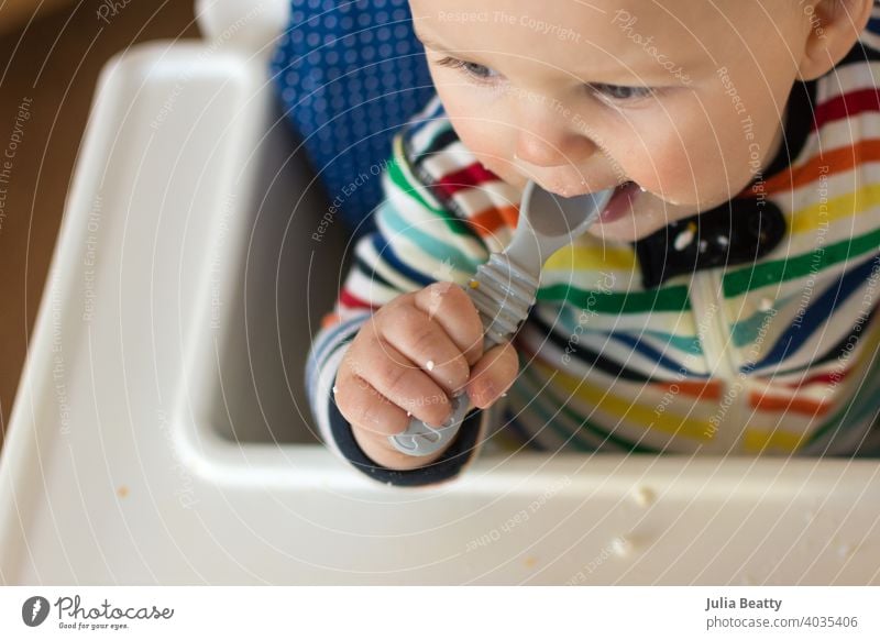 Junges Baby im Regenbogenstreifen-Outfit füttert sich selbst mit einem Silikonlöffel; babygeführte Entwöhnung Löffel mit dem Löffel gefüttert packen Säugling