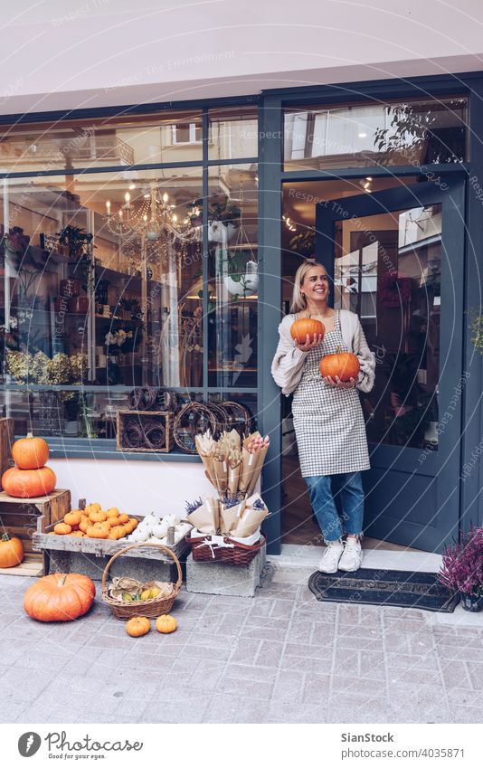 Lächelnde Frau hält Kürbisse vor ihrem Geschäft Glück Blumenhändler Werkstatt Konzept Hintergründe Gesundheit Laden Halt Mädchen Schürze orange urban im Freien