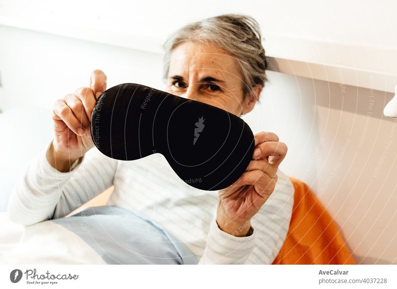 Eine alte Dame zeigt eine Schlafmaske in die Kamera, während sie in einem modernen Schlafzimmer lächelt Mundschutz Frau Senior Nacht schlafen Gesicht grauhaarig