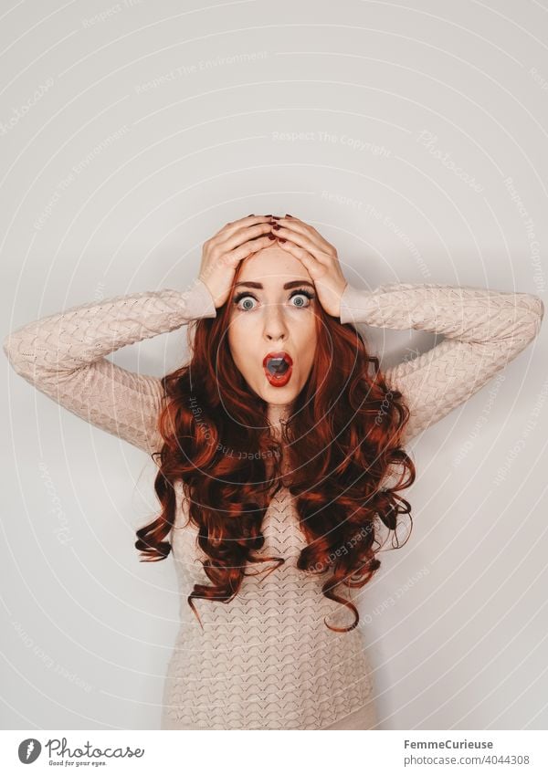Frau mit langen roten lockigen Haaren und erschrockenem Gesichtsausdruck schlägt beide Hände über dem Kopf zusammen schockiert überrascht Neutraler Hintergrund
