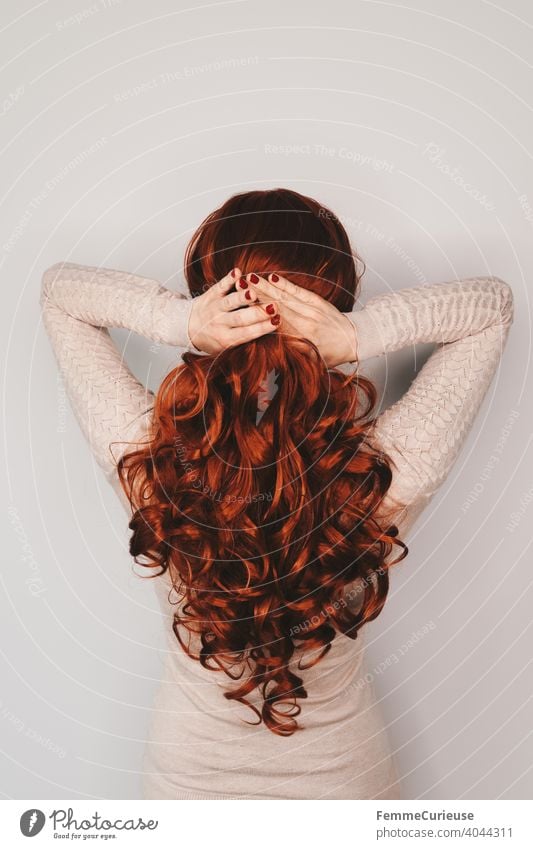 Rückansicht einer Frau mit langen roten lockigen Haaren in hautfarbenem Pullover die ihre Haare mit beiden Händen zusammenhält zusammenhalten festhalten