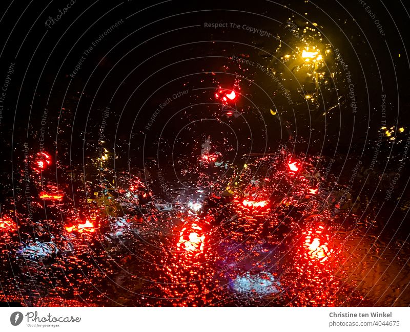 Blick durch die verregnete  Windschutzscheibe eines Autos, das in der Dunkelheit hinter beleuchteten Fahrzeugen an einer roten Ampel steht Lichter