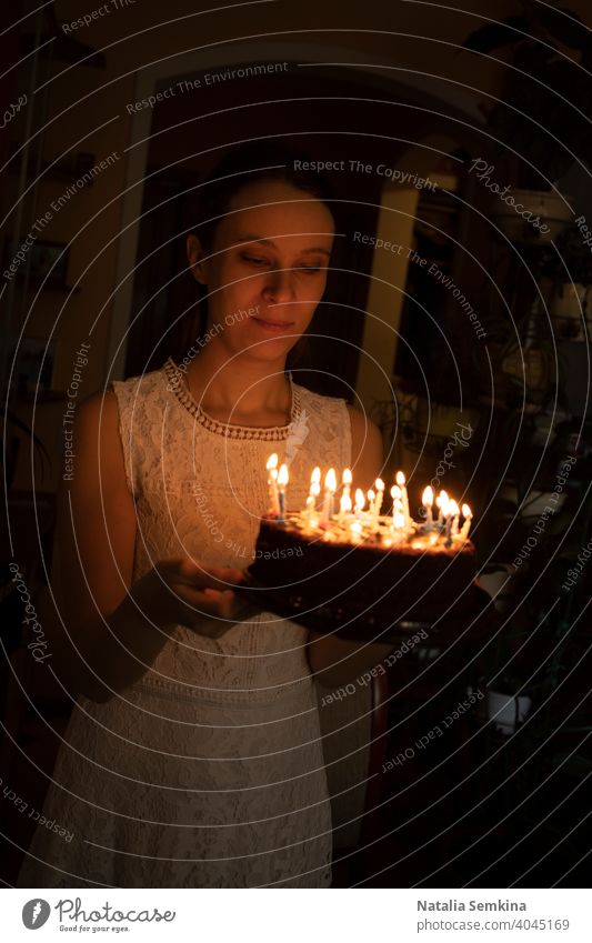 Junges Mädchen in weißem Spitzenkleid hält Kuchen mit brennenden Kerzen in ihren Händen in einem dunklen Raum. Geburtstagsfeier zu Hause. Vertikale Ausrichtung.