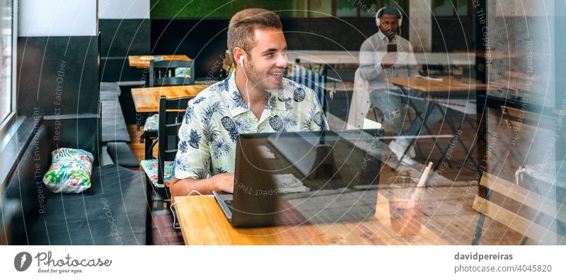 Junger Mann mit Kopfhörern und Laptop in einem Kaffeehaus Panorama Transparente Glück Freizeitkleidung zur Seite schauend kostenloses W-LAN sprechend