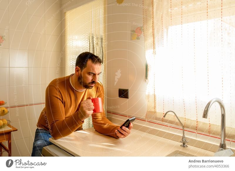 Hübscher bärtiger Mann im orangefarbenen Pullover, der in der Küche bei einer Tasse Kaffee auf das Smartphone schaut Handy Aussehen Mobile Mitteilung Telefon
