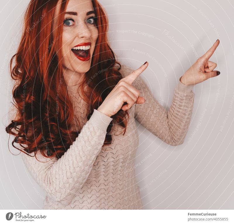 Frau mit langen roten lockigen Haaren zeigt mit beiden Händen und Zeigefingern auf etwas und lacht dabei in die Kamera Hinweis hinweisend zeigen hinzeigen